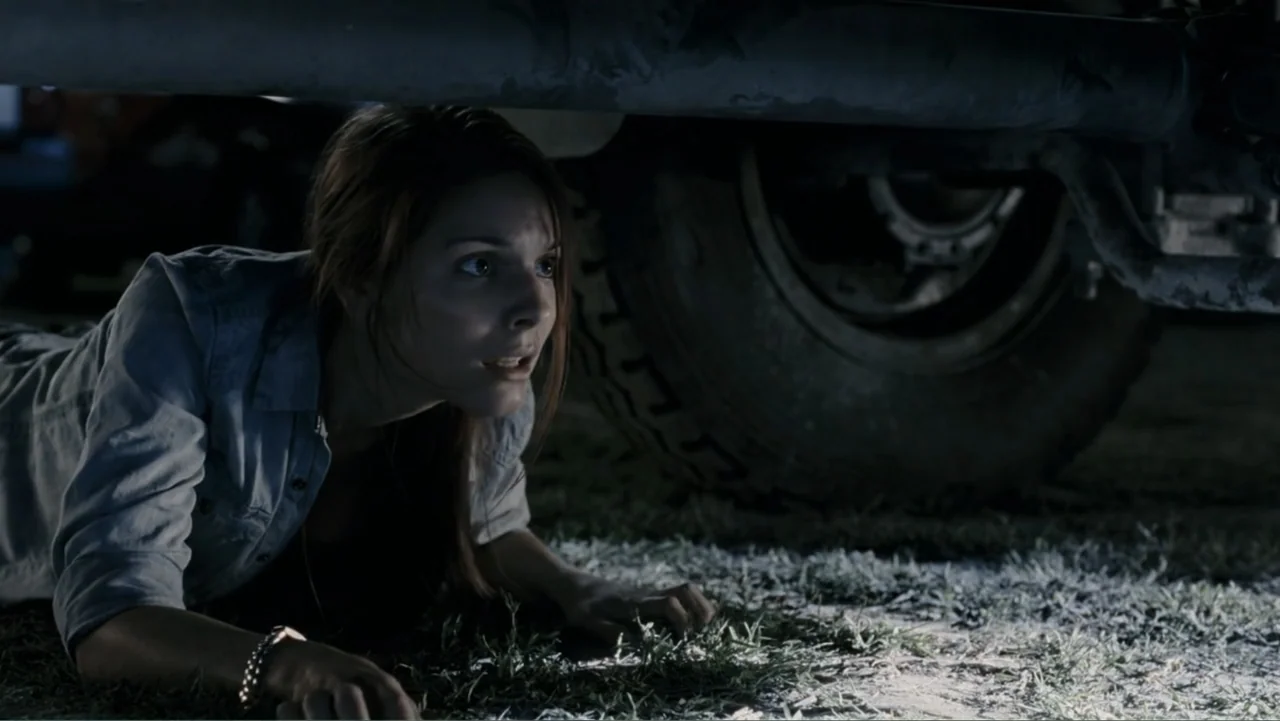 Tomorrow, When the War Began (2010) movie still: medium — A woman lies on the ground under the chassis of a large vehicle at night. She gazes ahead…