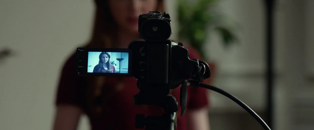 Oculus (2013) movie still: medium — A woman stands behind a tripod-mounted camera in a modern interior setting. The camera's flip-out screen…