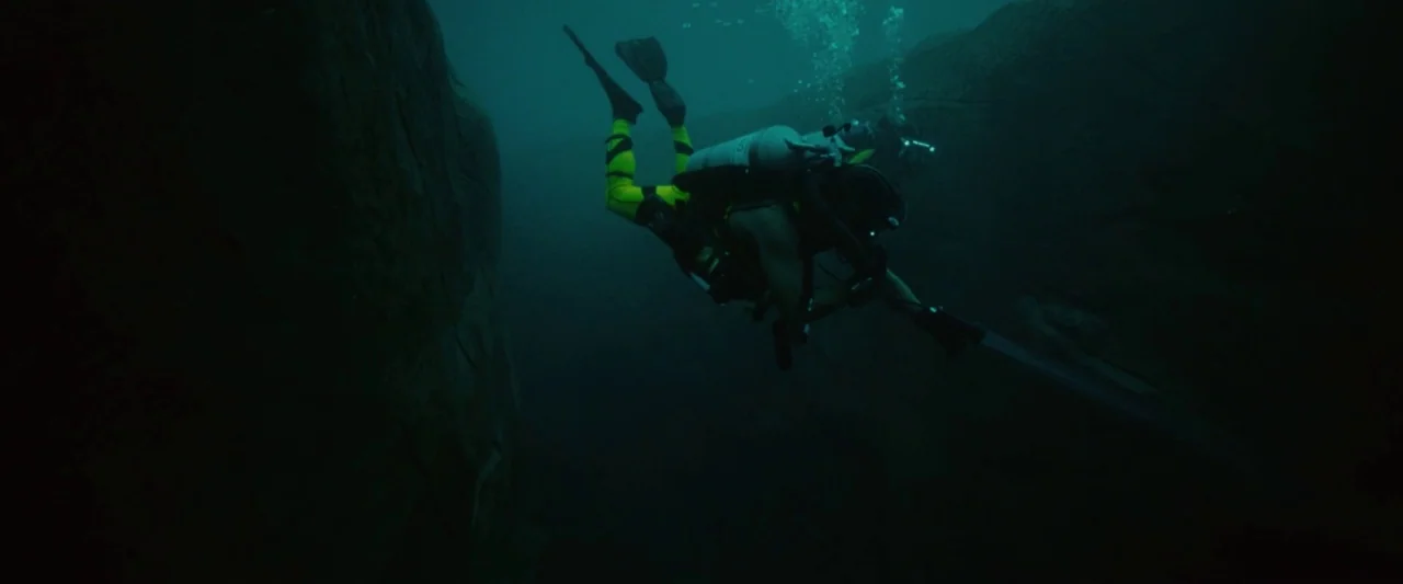 Piranha 3D (2010) movie still: wide — A dark underwater rock canyon with steep jagged stone walls on both sides. A scuba diver wearing a bright…