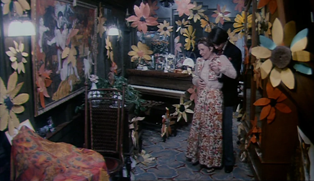Harold and Maude (1971) movie still: medium — A cluttered room features walls adorned with large hand-cut paper flowers and a painting. A couple…
