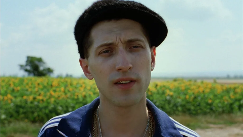 Everything Is Illuminated (2005) movie still: closeup — A field of sunflowers sits under an overcast sky during the day. A young man wearing a hat…