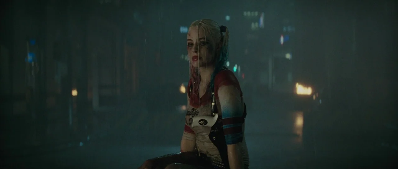 Suicide Squad (2016) movie still: medium — A dark, rainy urban street at night with distant faint glowing lights. A blonde woman with disheveled…