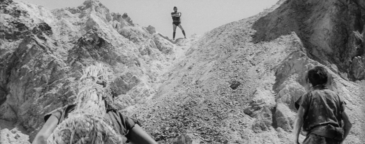 The Hidden Fortress (1958) movie still: wide — A rugged quarry with steep, light-colored dirt and rock slopes under bright light. A man stands alone…