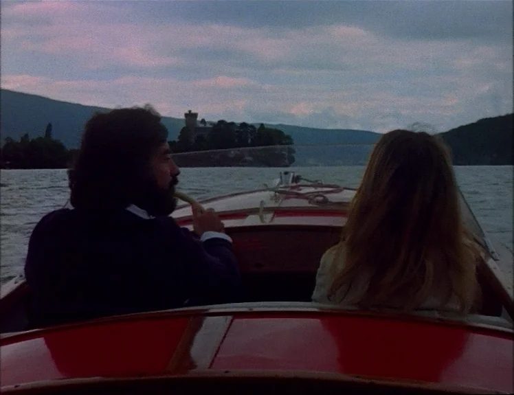 Claire’s Knee (1970) movie still: medium — A man and woman travel in a red motorboat on a calm, overcast lake. Distant mountains and a small castle…