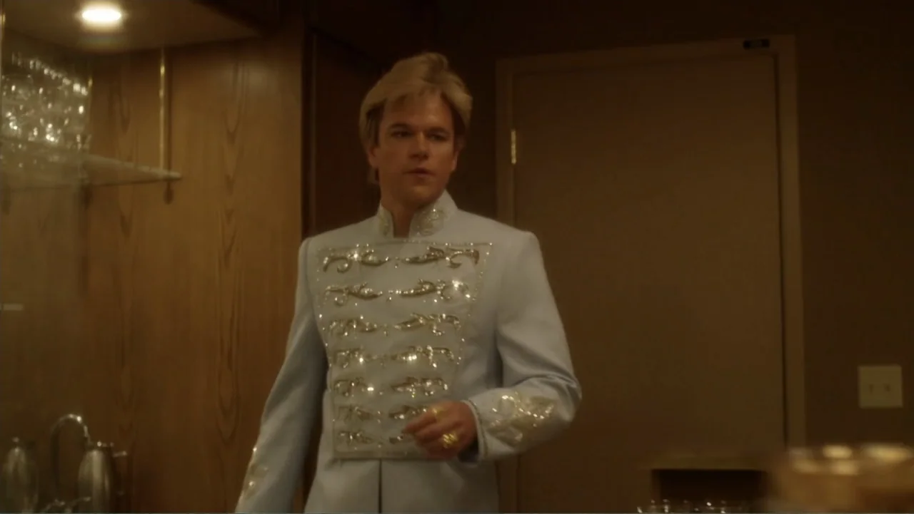 Behind the Candelabra (2013) movie still: medium — A dimly lit room with wood-paneled walls features a single light source. A man wearing a light…