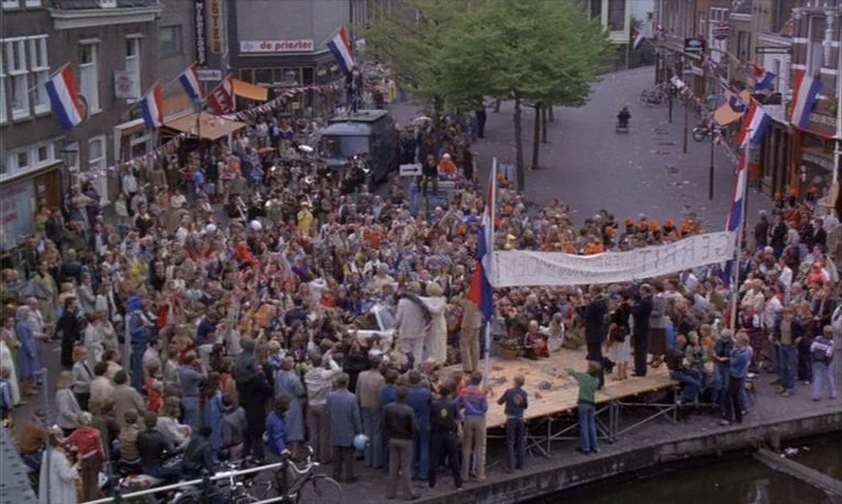Spetters (1980) movie still: wide — A dense crowd fills a European city street lined with brick buildings and flags. People gather around a wooden…