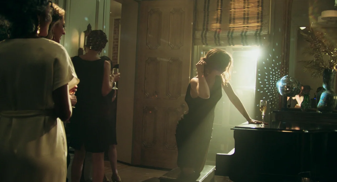 Deep Water (2022) movie still: medium — A dimly lit room with people gathered at a party. A woman in a black dress leans on a grand piano near a…
