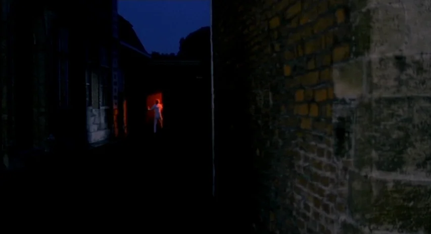Malpertuis (1971) movie still: wide — A dark, narrow outdoor passageway is flanked by high stone and brick walls at night. A person in a light suit…