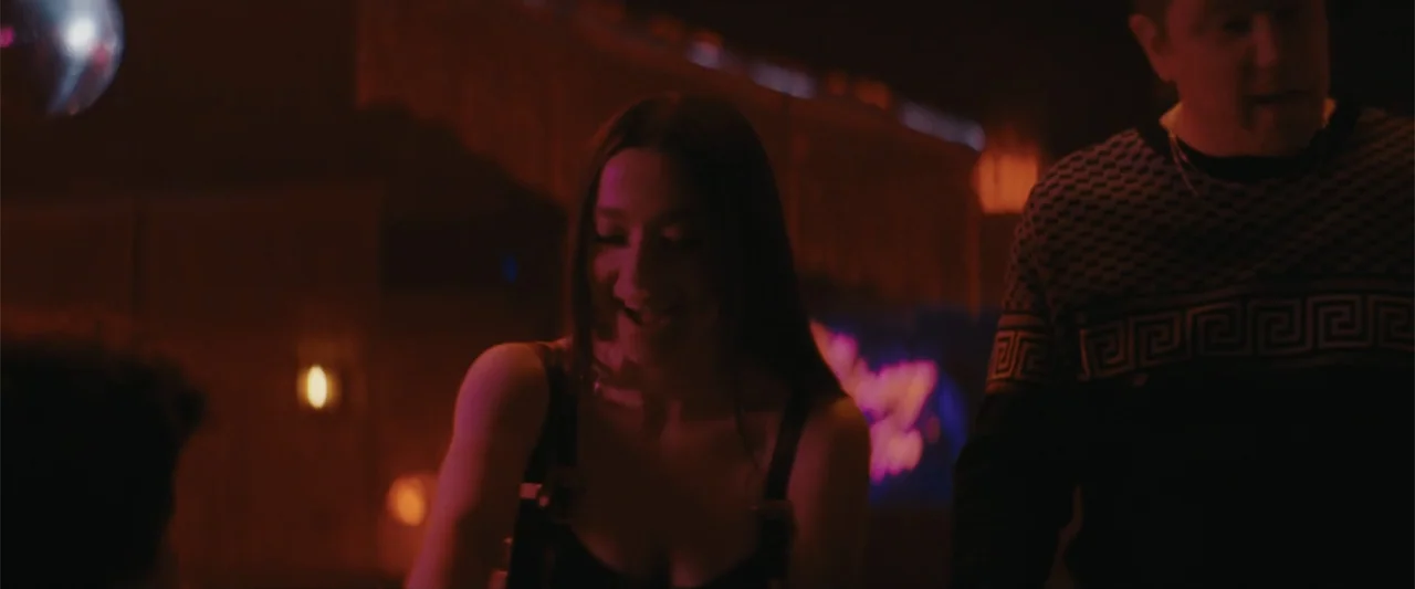 Anora (2024) movie still: medium — A dimly lit club interior features dark surfaces and vibrant magenta neon lighting. A young woman and a man in a…