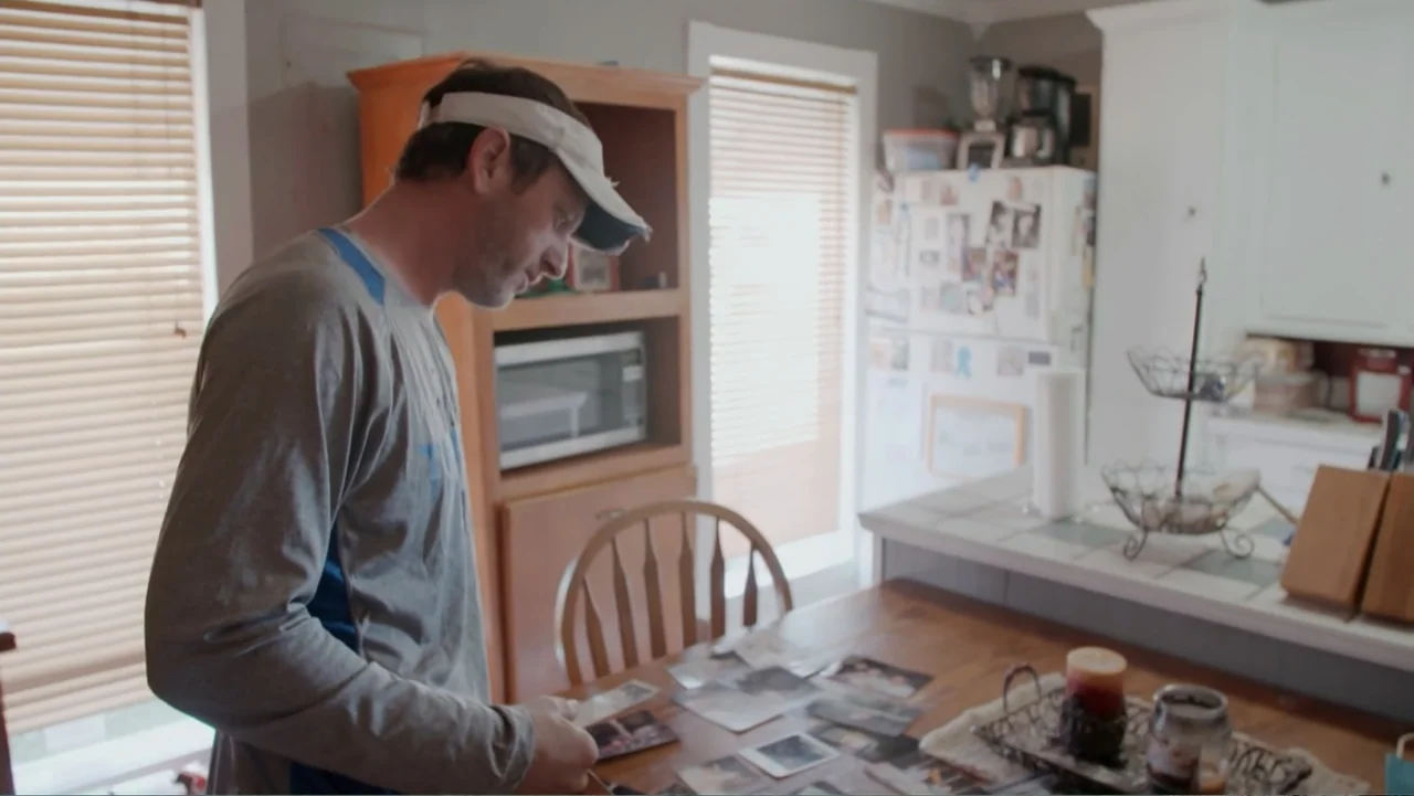 Mommy Dead and Dearest (2017) movie still: medium — A dining area contains a wooden table scattered with several rectangular photographs. A man…
