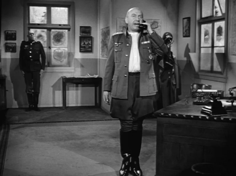 Stalag 17 (1953) movie still: fullbody — A military office features maps on the wall and two desks under large windows. A bald officer stands in the…