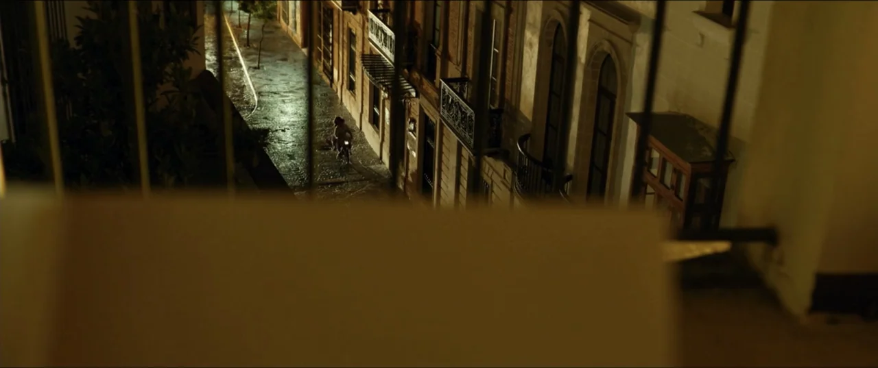 Hear Me: Our Summer (2024) movie still: establishing — A narrow stone alleyway is viewed from a high balcony behind iron railings. A single wet…