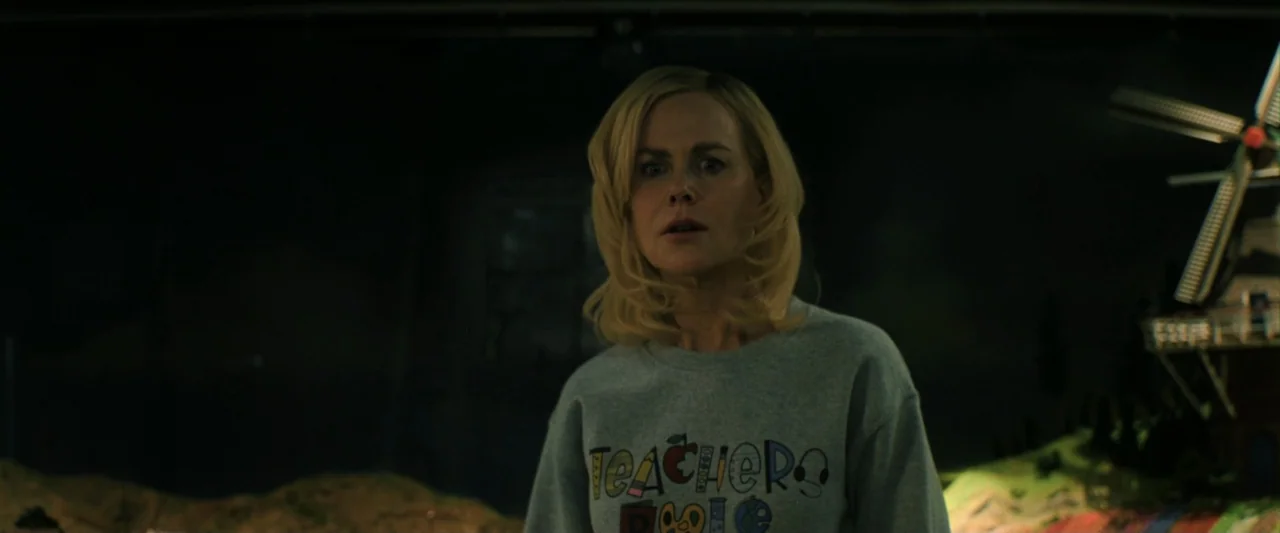 Holland (2025) movie still: medium — A dimly lit room features a model windmill in the dark background. A woman in a gray sweatshirt looks directly…