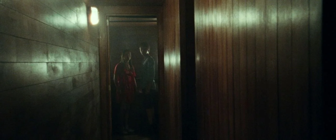 Adoration (2019) movie still: wide — A dark hallway with wood-paneled walls features a bright doorway at the far end. A young woman and a young man…