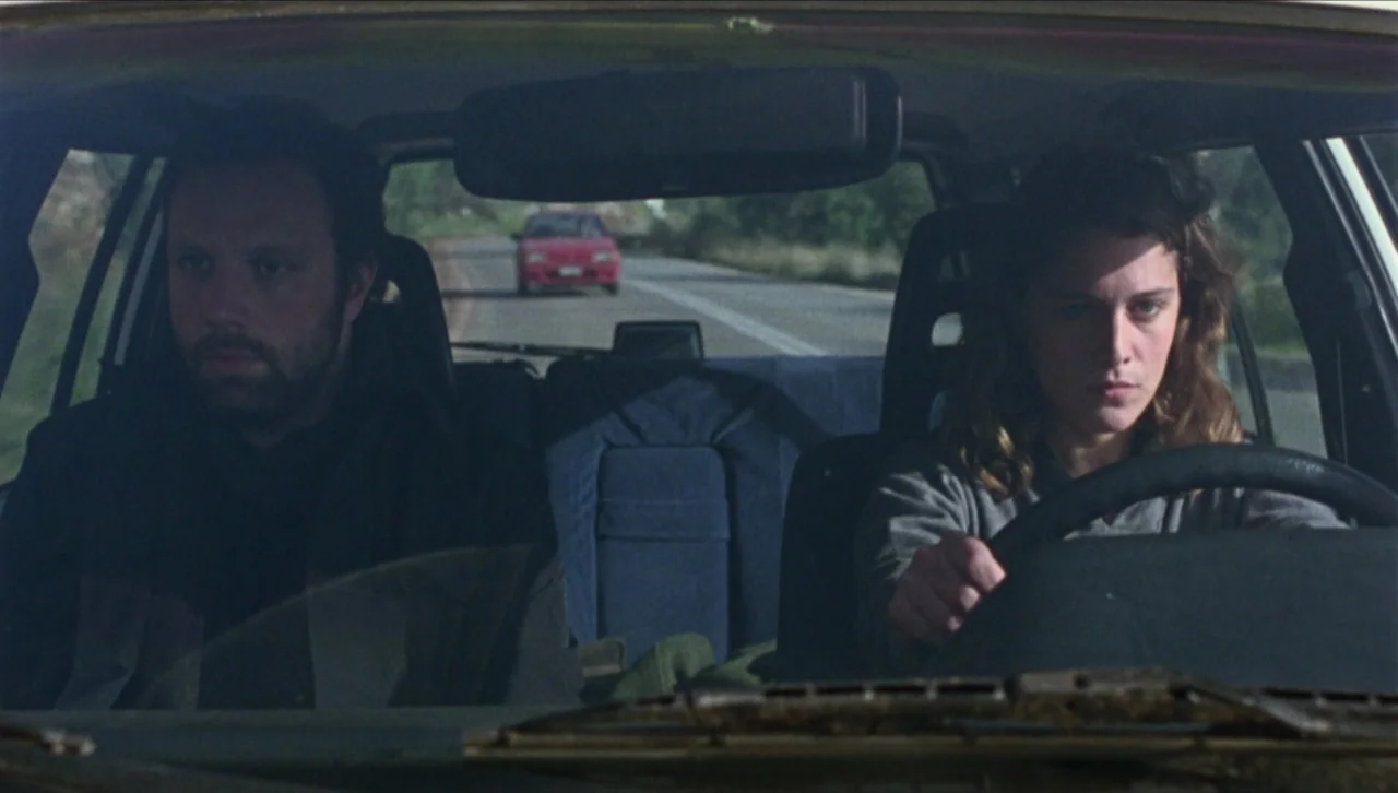 Attenberg (2010) movie still: medium — A man and woman sit in the front seats of a vehicle driving on a road. A red car travels on the road in the…