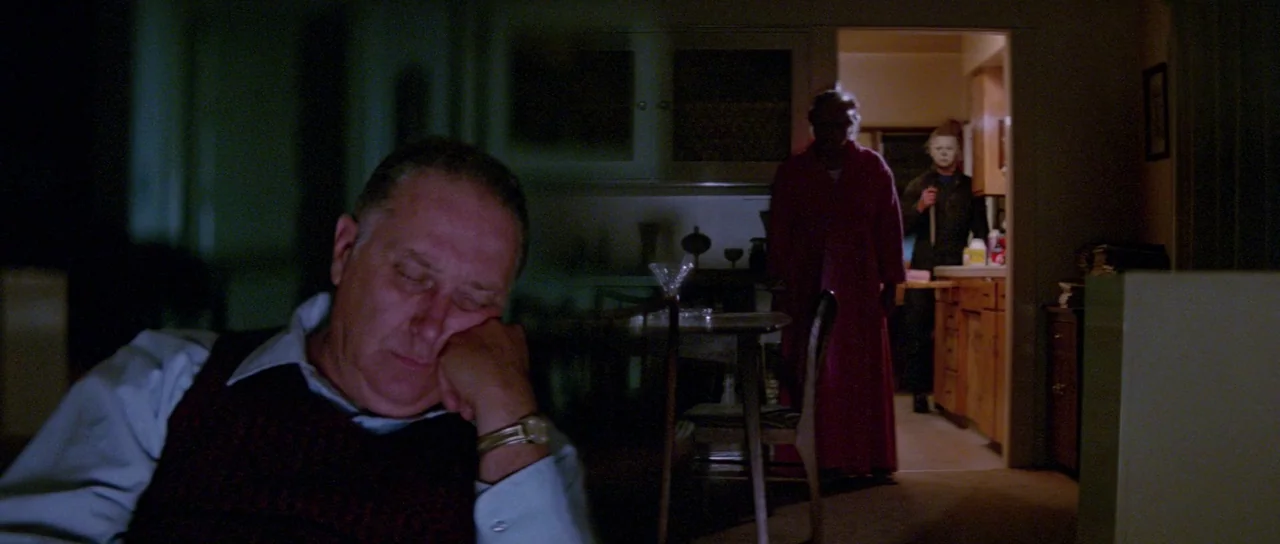 Halloween II (1981) movie still: medium — A dimly lit room with an older man sleeping in a chair at the foreground. In the distant doorway, a masked…