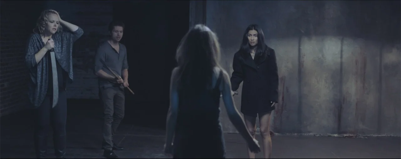 El Último Juego (2025) movie still: wide — An empty room with bare concrete walls stained with vertical streaks of dark red. Four people face each…