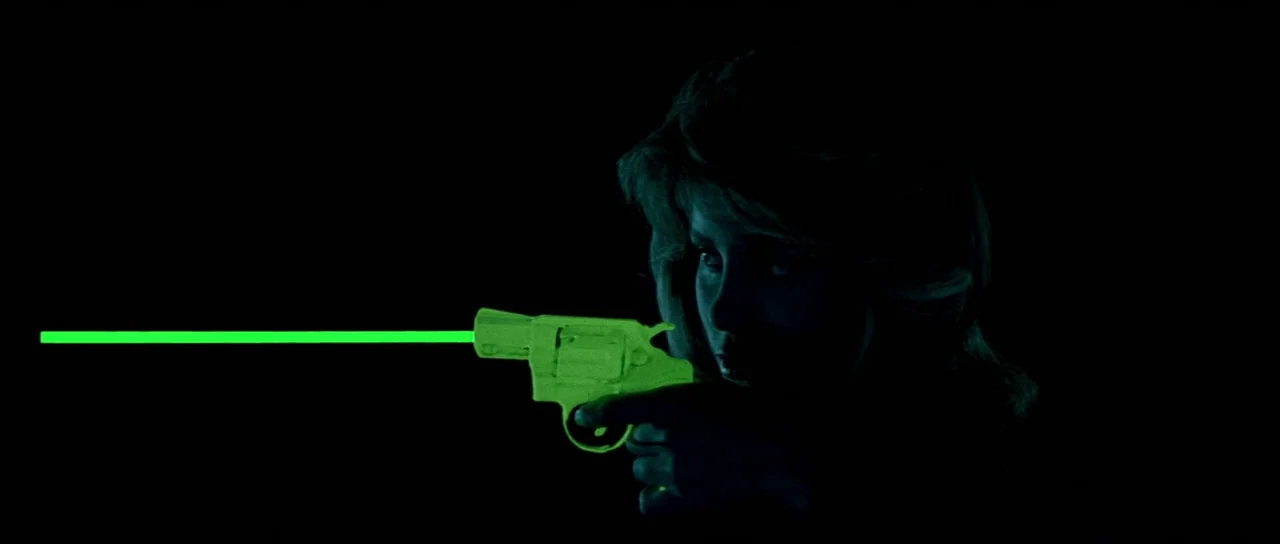 A View to a Kill (1985) movie still: closeup — A woman stands in a completely dark environment. She aims a brightly glowing lime-green revolver…