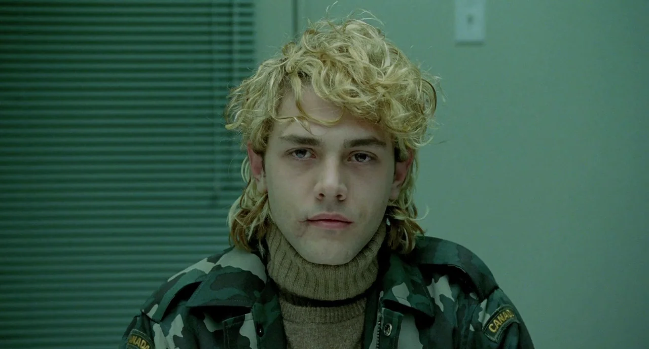 Tom at the Farm (2013) movie still: closeup — An interior space with pale green walls and horizontal window blinds behind a subject. A young man…