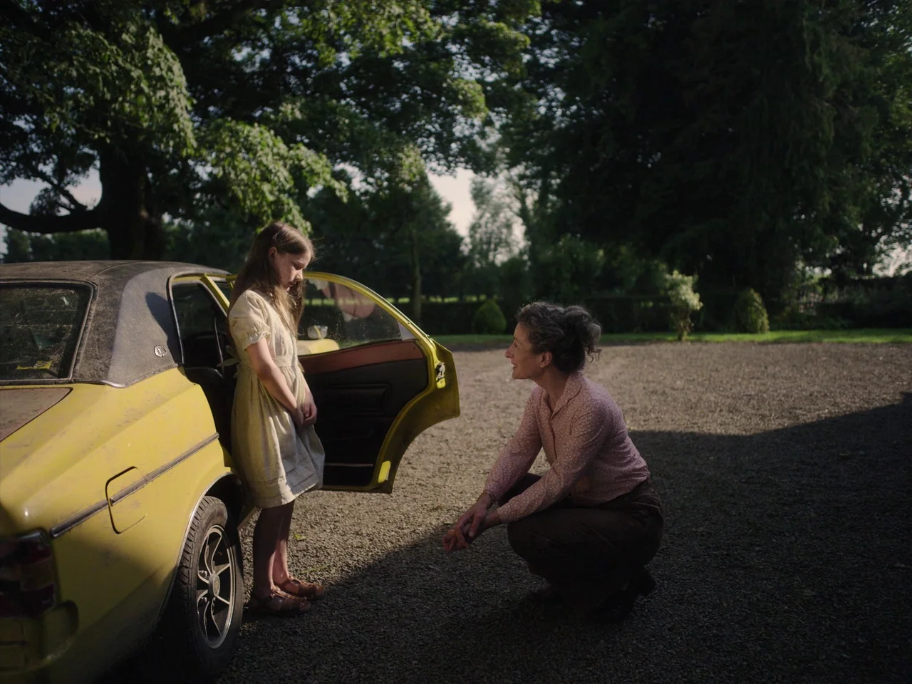 The Quiet Girl (2022) movie still: medium — A gravel driveway sits beneath large trees in the bright afternoon sunlight. A woman kneels beside an…