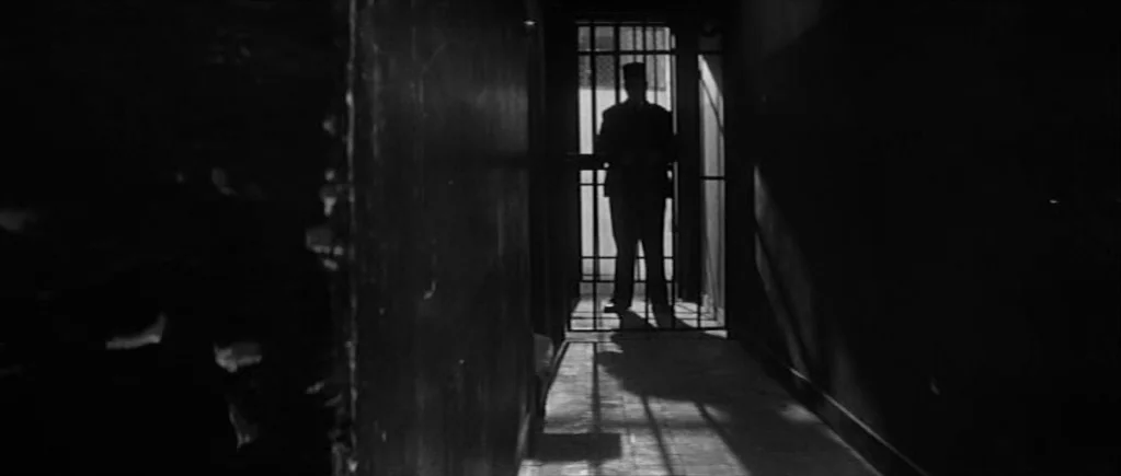The 400 Blows (1959) movie still: wide — A dark, narrow passageway features a gated entrance at the end of the hall. A man in a uniform stands…