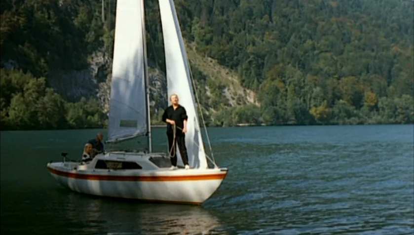 Funny Games (1997) movie still: wide — A small sailboat floats on a calm lake bordered by a dense, tree-covered hill. A woman in black clothing…