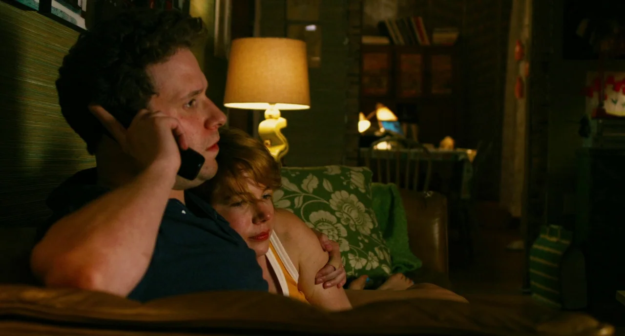 Take This Waltz (2011) movie still: medium — A couple sits closely on a sofa in a dimly lit, cluttered home space. The man holds a black smartphone…