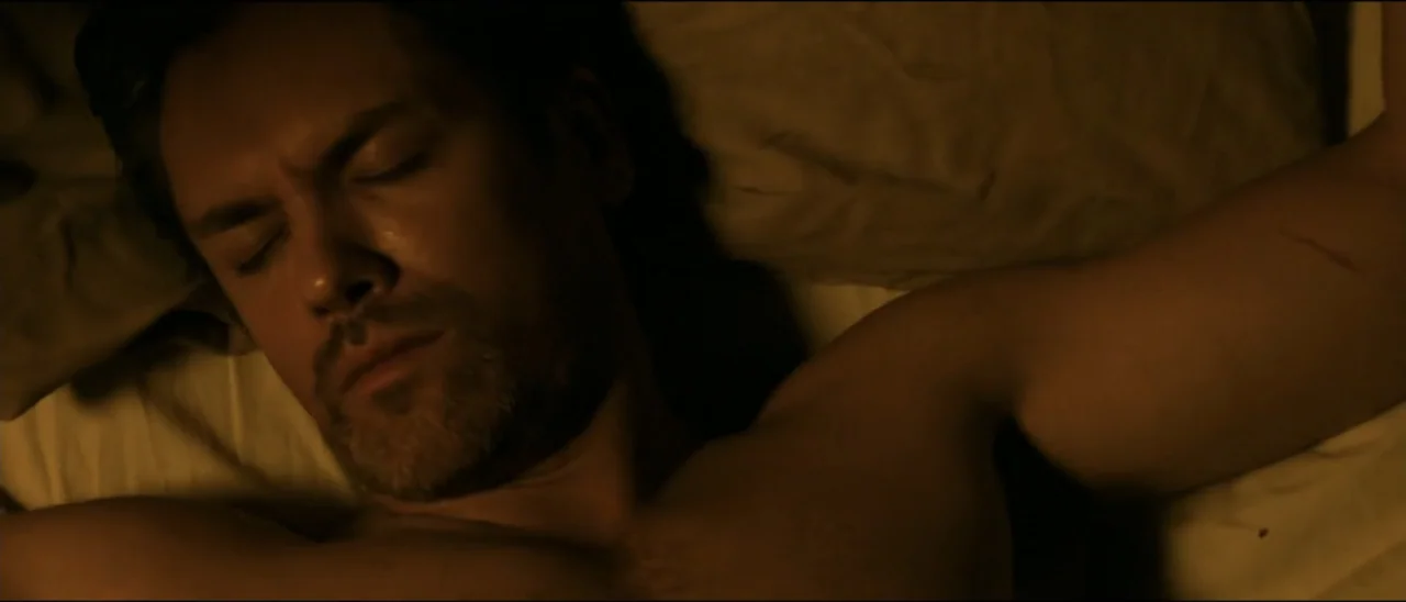 Dead for a Dollar (2022) movie still: closeup — A close-up of a man lying in a bed with dark linens and pillows. The man has his eyes closed and is…