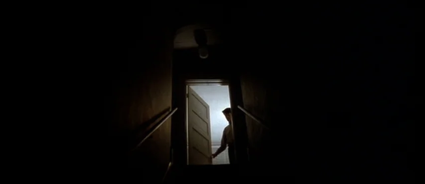 Blue Velvet (1986) movie still: wide — A dark stairwell with metallic handrails leads to an illuminated doorway at the top. A man stands in the…