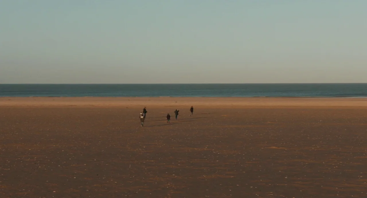 Everyday (2012) movie still: wide — A vast, empty beach extends to the horizon under a clear, pale sky. Four distant figures in dark clothing walk…