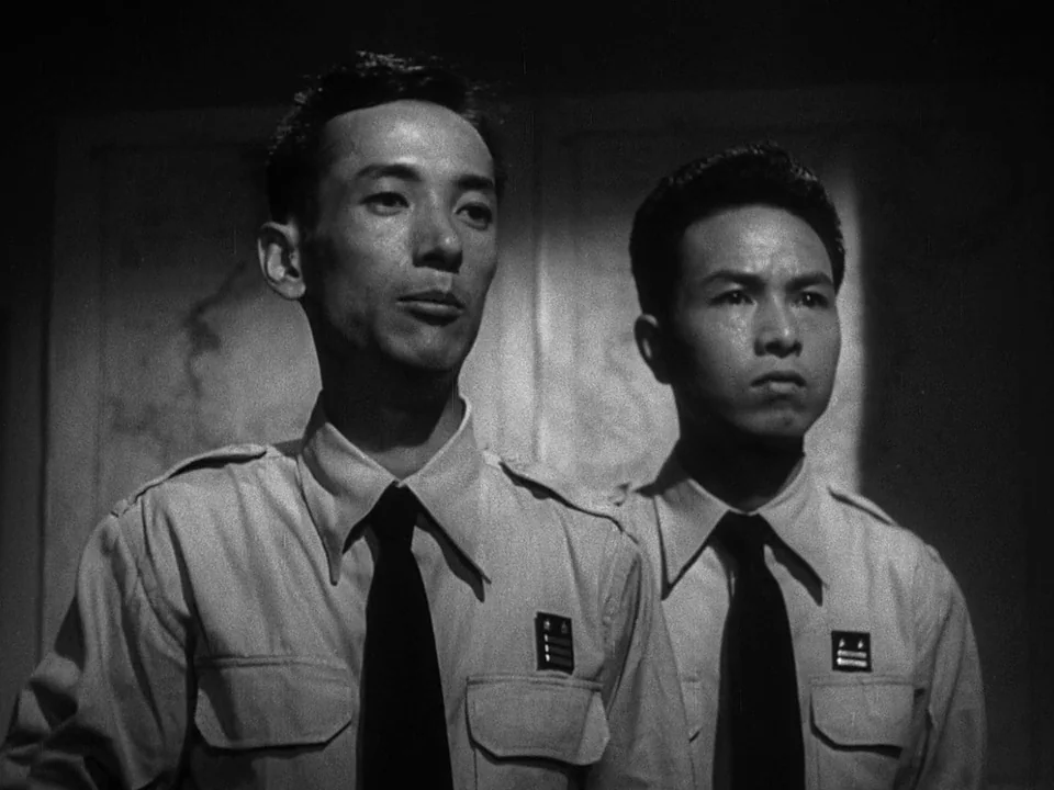 Godzilla (1954) (1954) movie still: medium — A dimly lit room features a plain wall background behind two uniformed men. The men stand perfectly…