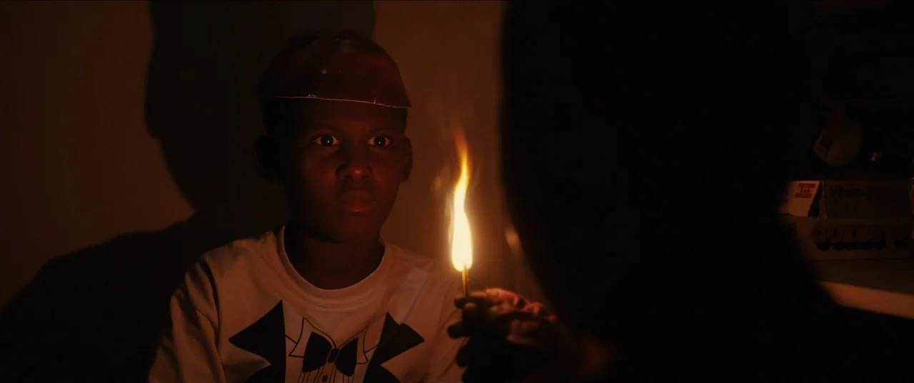 Us (2019) movie still: medium — A dimly lit interior space features a young boy watching a flickering match flame. The boy holds a small, flat…