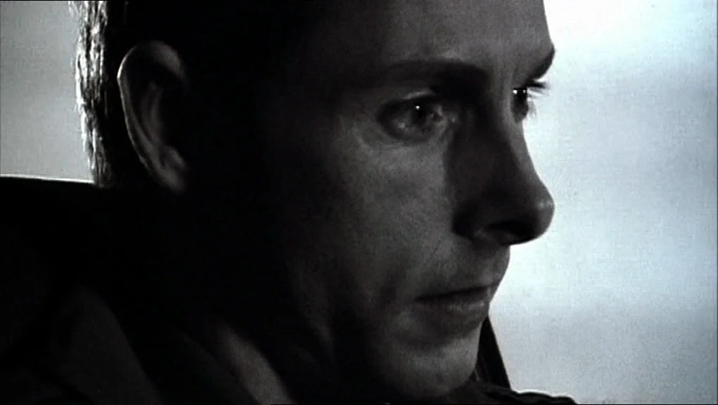 24 Hour Party People movie still: closeup — A man sits in the driver seat of a vehicle at night. The man gazes forward through the windshield with a…