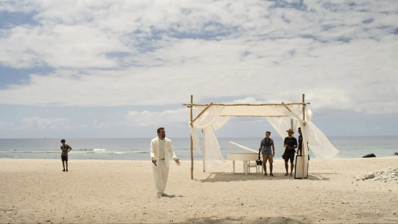 Unknown #1 movie still: wide — A sunny beach features a white piano under a bamboo canopy with fabric drapes. A man in a cream suit holds a pistol…
