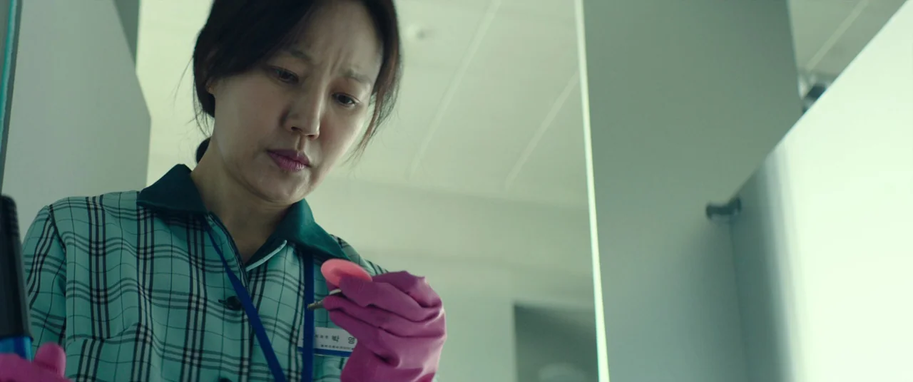 Beasts Clawing At Straws (2020) movie still: medium — A sterile public restroom with white tiled walls and fluorescent lighting. A woman in work…