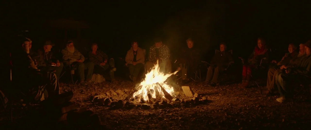 Nomadland (2020) movie still: wide — A group of people are gathered at night around a bright, crackling campfire outdoors. They sit on folding…