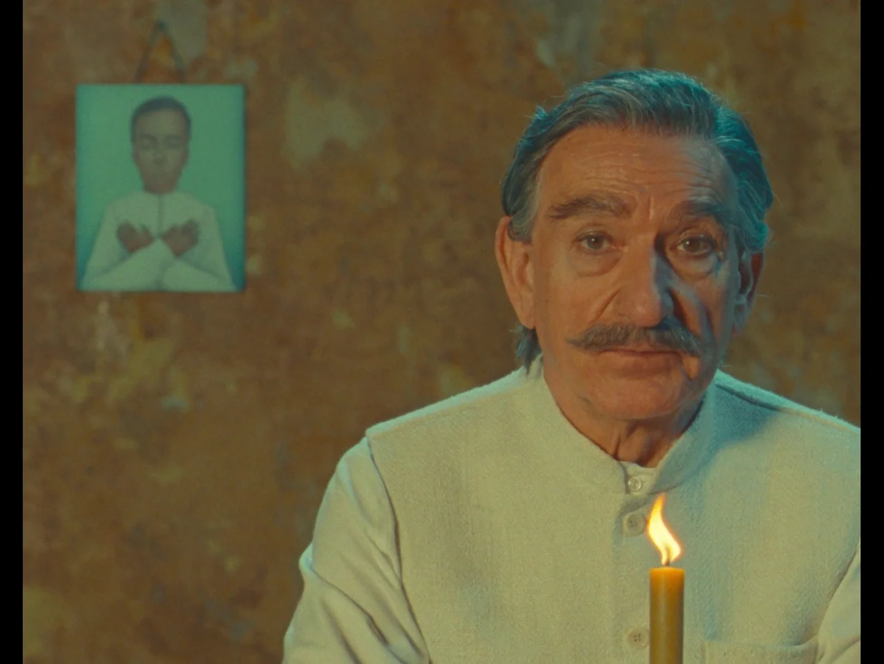 The Wonderful Story of Henry Sugar (2023) movie still: medium — An aged man sits in a dimly lit room against a weathered, yellow-toned wall. He…