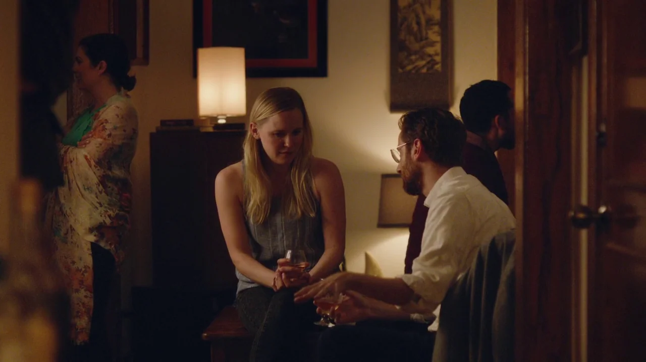 Saint Frances (2019) movie still: medium — An interior space with warm lighting and framed artwork on the beige walls. A man and woman sit facing…