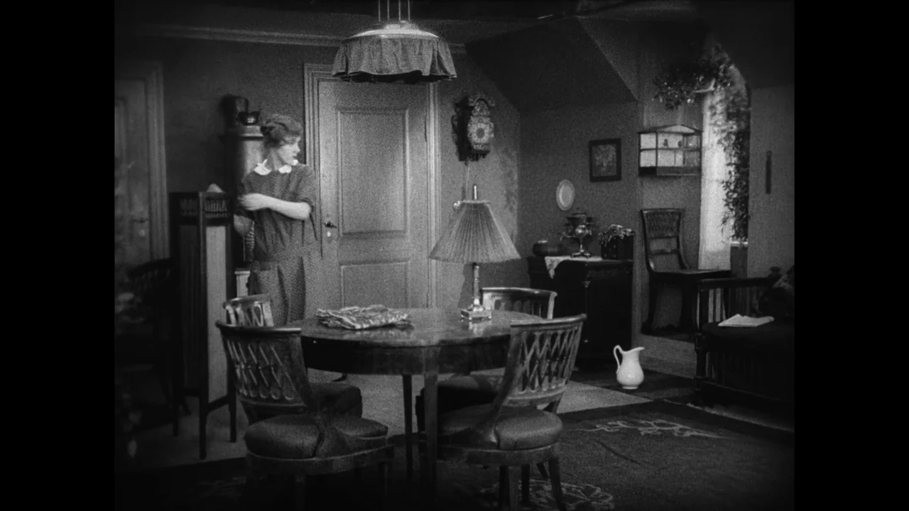 Master of the House (1925) movie still: wide — A 1920s interior featuring a round wooden table, chairs, a wall clock, and lamp. A young woman in a…