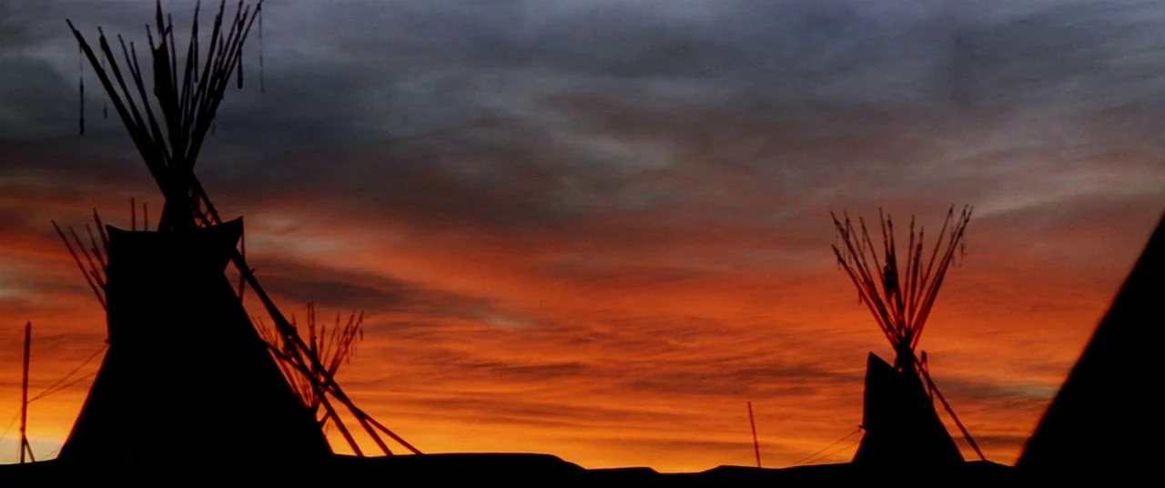Dances With Wolves (1990) movie still: wide — Three teepees stand as silhouettes against a dark, dramatic sunset sky at dusk. The sky features bands…