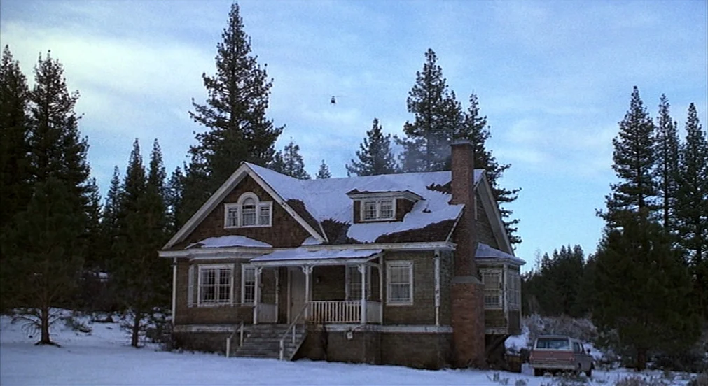 Misery (1990) movie still: wide — A weathered wooden house with snow-dusted roof sits amidst a grove of tall pine trees. A small helicopter appears…