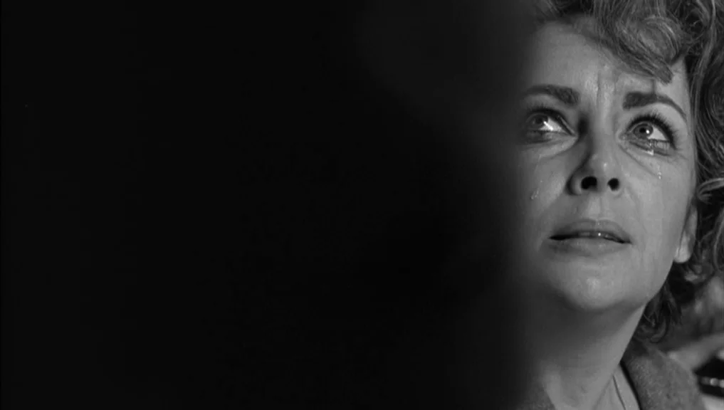 Who’s Afraid Of Virginia Woolf? (1966) movie still: closeup — A dark indoor space where a woman is partially obscured by a foreground element. The…