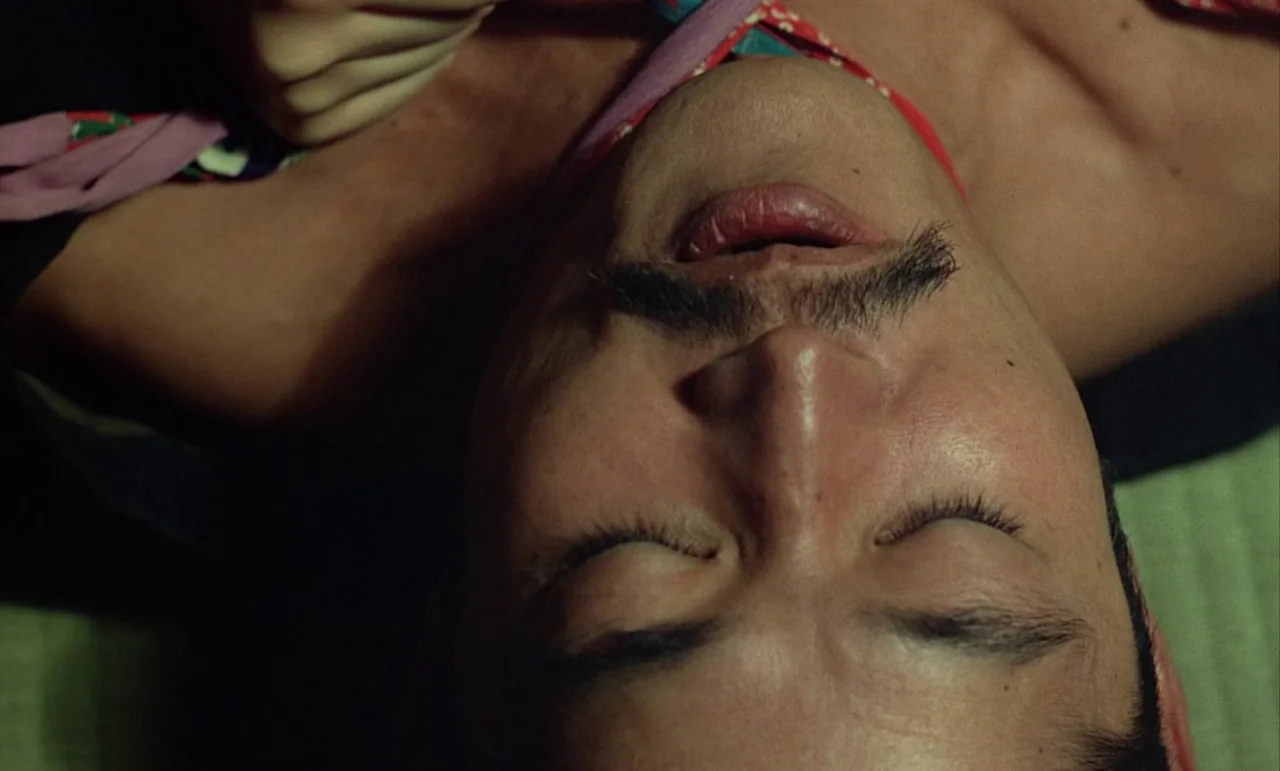In The Realm of the Senses (1976) movie still: closeup — A man lies on a woven straw mat with patterned textile accents. The camera is positioned…