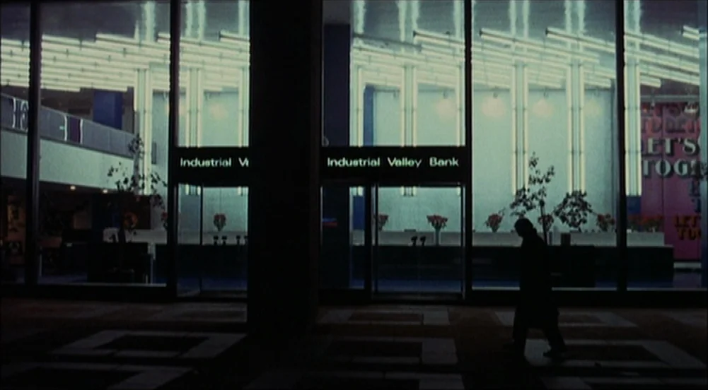 The King Of Marvin Gardens movie still: wide — A dark urban exterior features a bank storefront with large glass panels and neon signage. A…