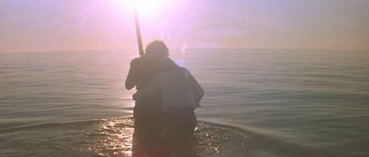 Mad Max (1979) movie still: medium — Two people stand together waist-deep in the calm water of the ocean. The sun sets directly behind them causing…