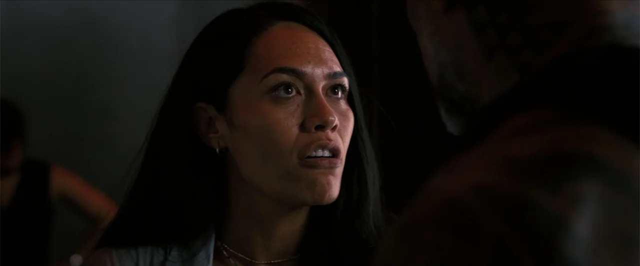 The Wrecking Crew (2026) movie still: closeup — A dimly lit interior space with shadows obscuring the background. A woman in her thirties looks…