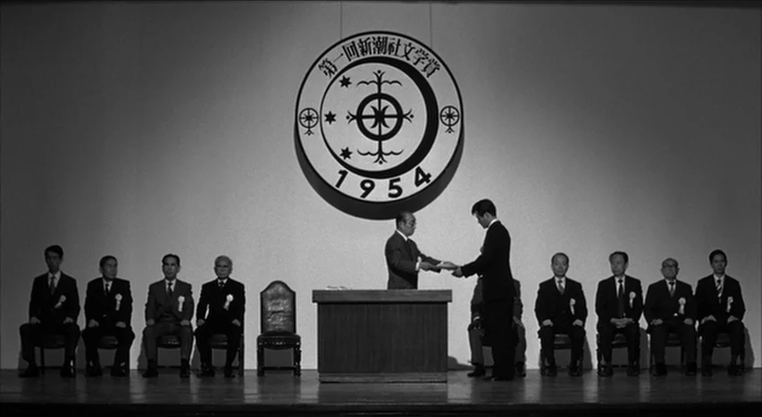 Mishima: A Life in Four Chapters (1985) movie still: wide — A formal awards ceremony takes place on a bare stage with a large emblem. A younger man…