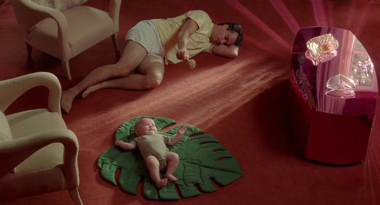 Babysitter (2022) movie still: wide — A spacious room with bright red carpeting and a large leaf-shaped mat on the floor. A man in yellow lies on…