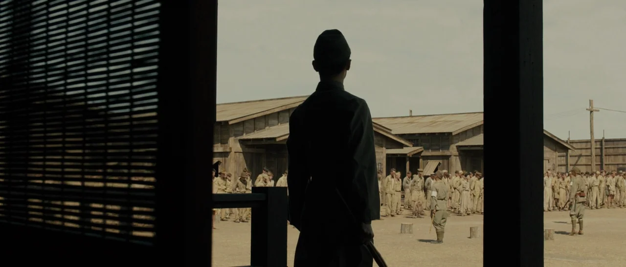 Unbroken (2014) movie still: medium — An outdoor military camp features wooden barrack structures surrounding a dry, dusty yard. A man in a dark…