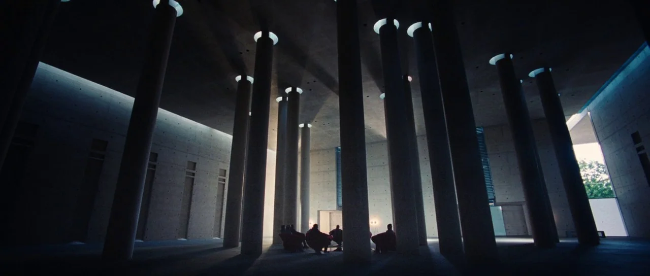 Aeon Flux (2005) movie still: wide — A vast hall constructed of smooth concrete columns rises to a high ceiling with skylights. Several people sit…